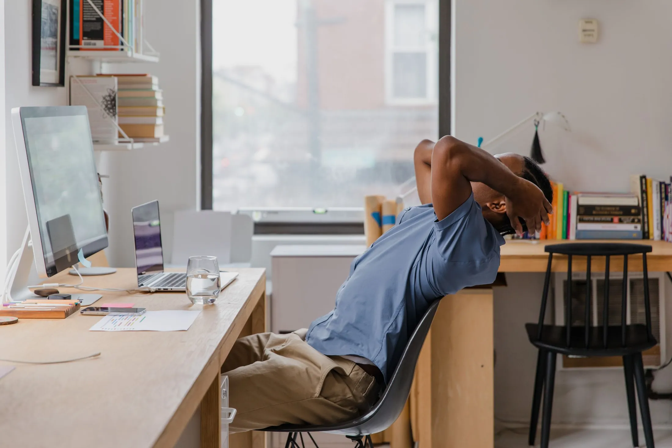 A man at a home-office desk leans back in his chair with both hands clasped behind his neck, stretching beside a large window.
