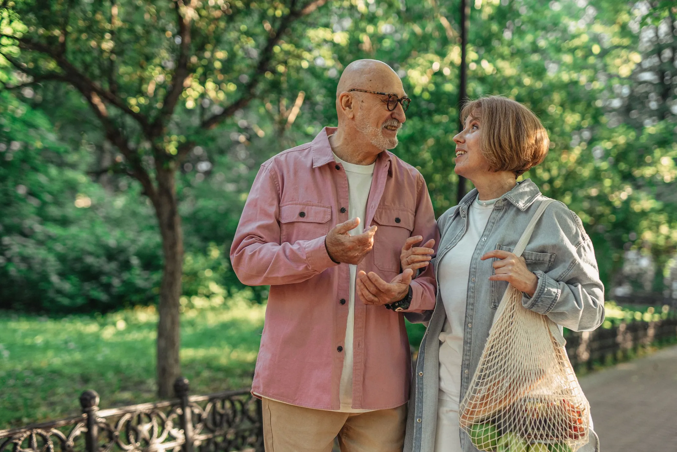 An older couple chat and laugh as they walk along a tree-lined neighbourhood path, the woman carrying a net bag of groceries.
