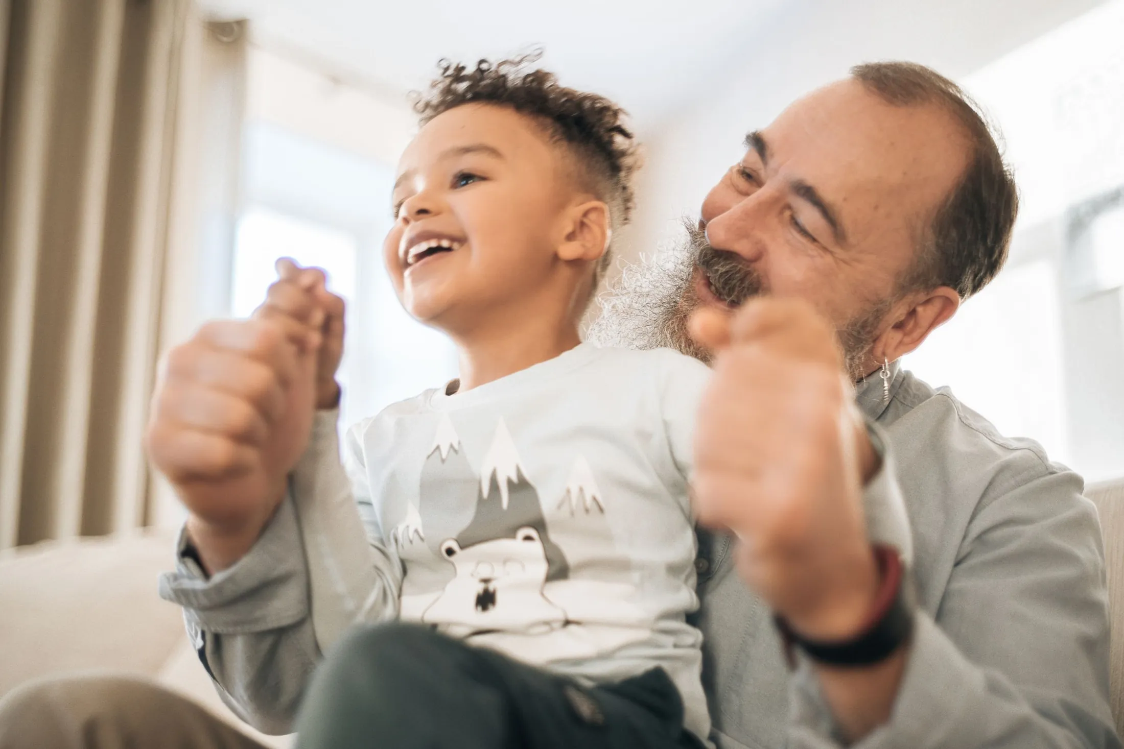 A laughing little boy sits on his bearded grandfather's lap at home as they hold hands and play.