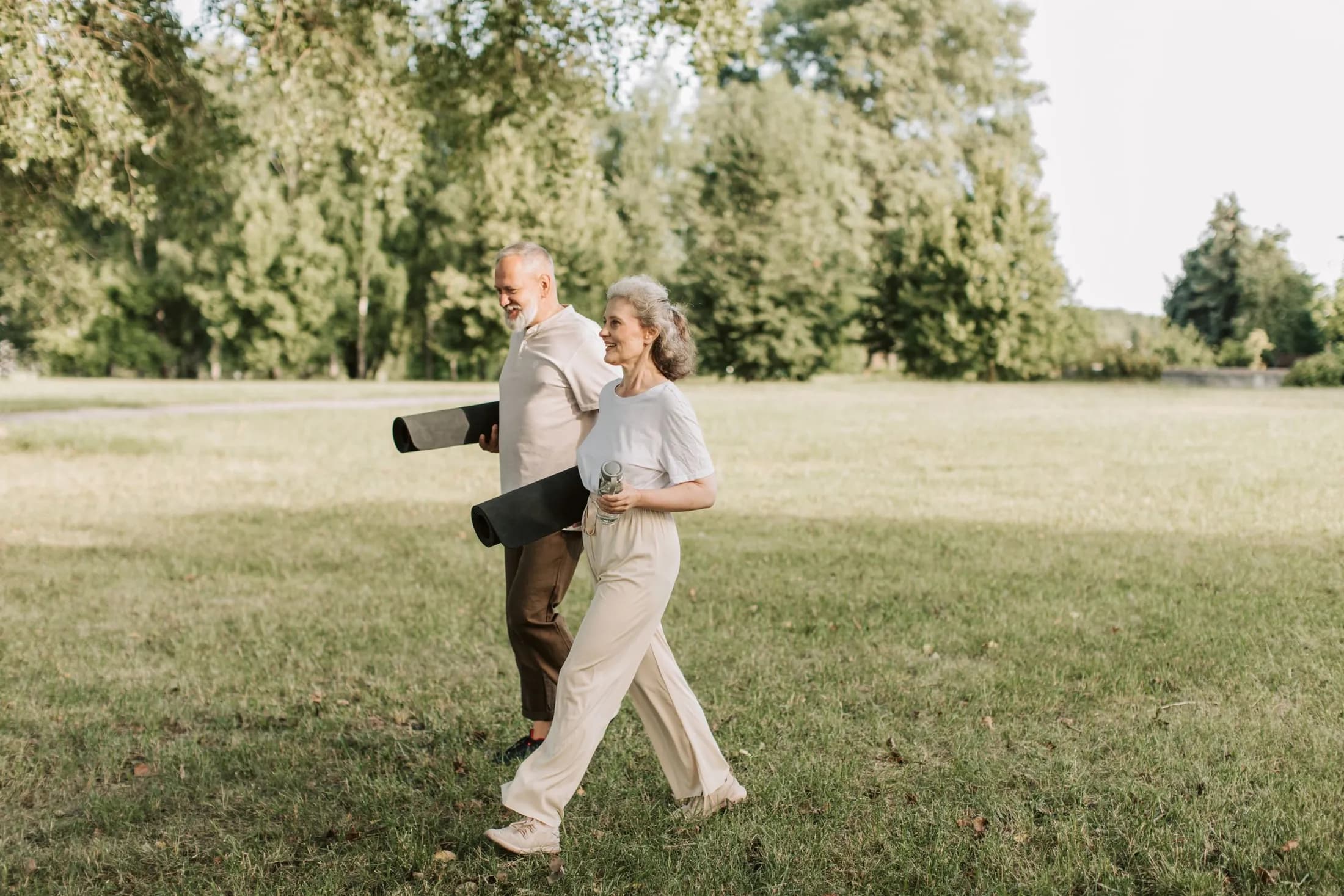 An older couple with grey hair walk side by side across a sunlit park lawn carrying rolled exercise mats, smiling and looking ahead.