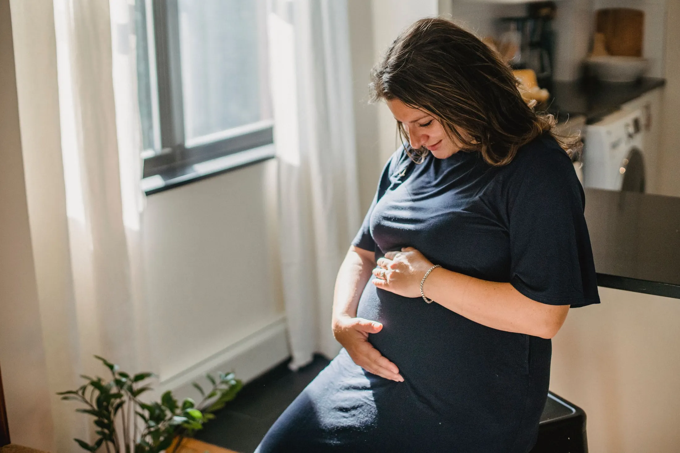 A pregnant woman in a navy dress sits by a sunlit kitchen window, smiling down with her hands resting on her belly.