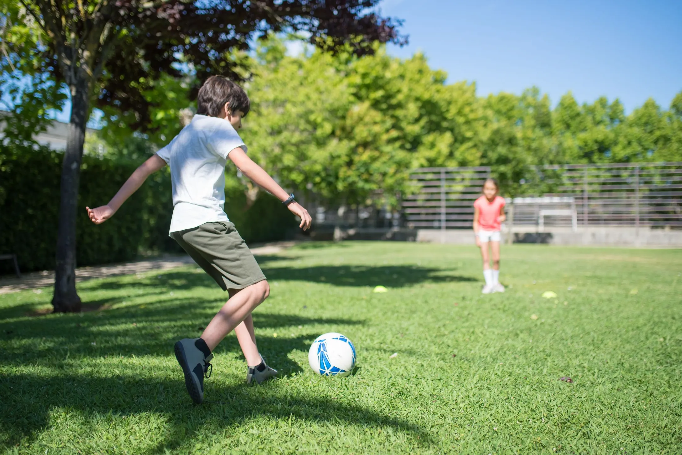 A boy swings his leg to kick a soccer ball across a sunny park lawn toward a girl waiting in the distance.